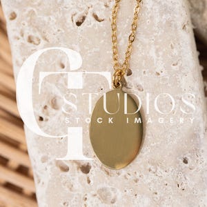 May include: A gold-colored oval pendant necklace. The pendant hangs from a delicate chain. The necklace is displayed on a textured, light-colored stone. The image includes the text "STUDIOS STOCK IMAGERY".