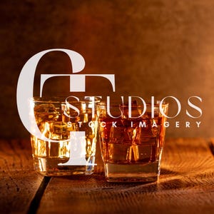 Puede incluir: Dos vasos cuadrados de whisky llenos de líquido ámbar y cubitos de hielo sobre una superficie de madera. Los vasos están iluminados, con un brillo cálido que resalta el contenido. La imagen tiene el texto "STUDIOS STOCK IMAGERY".
