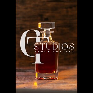 May include: A clear glass decanter with a square base and a stopper, filled with a dark amber liquid. The decanter is set against a wooden background. The text "STUDIOS STOCK IMAGERY" is visible.