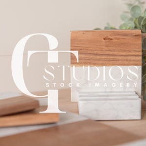 Puede incluir: Un soporte para teléfono de madera y mármol blanco con el texto "STUDIOS STOCK IMAGERY" en la parte delantera. El soporte está sobre una superficie marrón claro con una planta verde en el fondo.