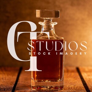 May include: A clear glass decanter filled with a dark amber liquid, likely liquor, sits on a wooden surface. The decanter has a square shape with a rectangular stopper. The background is a warm, blurred gradient.