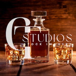 May include: A clear glass decanter with a square base and stopper, filled with amber liquid, stands between two lowball glasses filled with ice and amber liquid. The image is set against a wooden background.