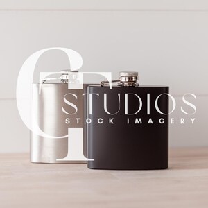 May include: Two stainless steel hip flasks, one silver and one black, on a white wood surface. The black flask has the text "STUDIOS STOCK IMAGERY" printed on it.