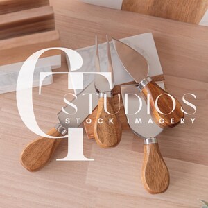 Puede incluir: Un juego de cuatro cuchillos para queso con mangos de madera. Los cuchillos están dispuestos en una tabla de cortar de mármol y madera.
