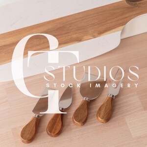 Puede incluir: Cuatro cuchillos de queso con mangos de madera y hojas de acero inoxidable sobre una superficie de madera. Los cuchillos están dispuestos en fila, con los mangos apuntando hacia el lado derecho de la imagen. La superficie es de color marrón claro, y hay una tabla de cortar blanca y marrón en el fondo.