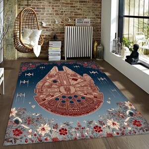 May include: A rectangular rug with a detailed illustration of the Millennium Falcon spaceship in brown and red against a blue gradient. The rug is bordered with floral patterns in red, white, and blue.