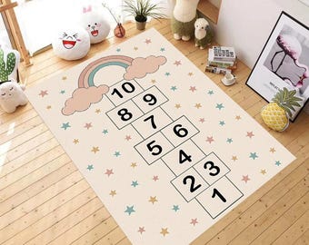 Puede incluir: Una alfombra beige de rayuela con un diseño de arcoíris y nubes. La alfombra presenta una cuadrícula de rayuela con los números del 1 al 10. Pequeñas estrellas de colores están dispersas por la alfombra. La alfombra está diseñada para niños.