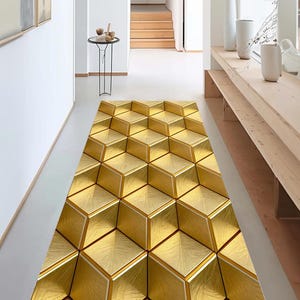 Può includere: Un tappeto runner con motivo geometrico color oro. Il tappeto presenta un motivo a cubi ripetuti con un aspetto strutturato. Il tappeto è posto in un corridoio con pareti bianche e pavimento chiaro.