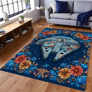 May include: A rectangular rug with a detailed image of the Millennium Falcon spaceship at its centre, framed by a floral border. The rug has a dark blue background with colourful flowers in shades of orange, yellow, and blue.