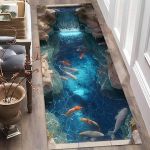 Op de afbeelding: Een rechthoekig tapijt met een gedetailleerd ontwerp van een koivijver. Het tapijt toont een waterval, rotsen en koi-vissen in oranje, wit en grijs tegen een blauwe waterachtergrond. Het tapijt ligt op een houten vloer.