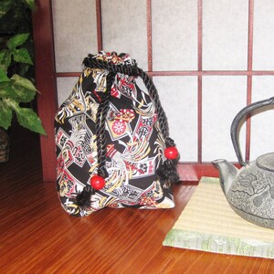 May include: A black drawstring bag with a Japanese-inspired design featuring red and gold accents. The bag has a black rope drawstring and red beads on the ends.