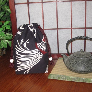 May include: A black drawstring bag with a white and red fan-like design. The bag has a burgundy drawstring and white ball accents. A cast iron teapot sits on a small mat next to the bag.