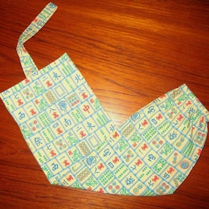 May include: A light yellow fabric bag with a handle, featuring a repeating pattern of colorful Mahjong tiles. The tiles are in shades of red, green, and blue, creating a playful and eye-catching design. The bag is open and laying on a wooden surface.