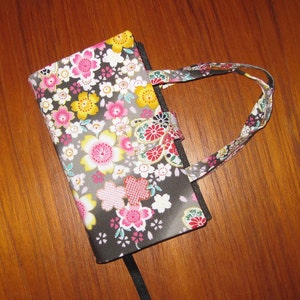 May include: A floral patterned book cover with a matching handle. The cover features pink, yellow, and white cherry blossoms on a gray and black background. The handle and closure have a similar floral design.