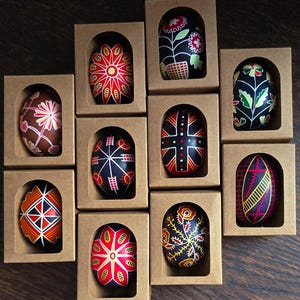 Puede incluir: Una colección de huevos de Pascua decorados con colores vibrantes, cada uno exhibido en cajas de cartón individuales. Los huevos presentan diseños intrincados en rojo, negro, amarillo y blanco, mostrando patrones florales y geométricos. Los huevos están sobre una superficie de madera marrón oscuro.