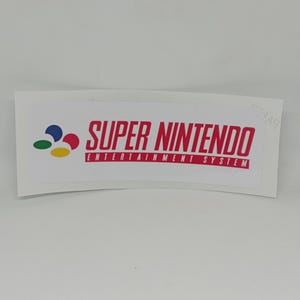 Puede incluir: Una pegatina blanca con el logotipo de Super Nintendo Entertainment System. El logotipo incluye las palabras "SUPER NINTENDO" en rojo, con "ENTERTAINMENT SYSTEM" debajo. La pegatina tiene un borde inferior curvo y un fondo blanco.