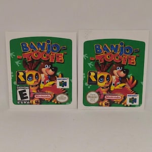 Op de afbeelding: Twee rechthoekige videogame-labels voor Banjo-Tooie met een beer, een vogel en een schedelfiguur op een groene achtergrond. De tekst Banjo-Tooie staat bovenaan. Eén label bevat een ESRB-beoordeling en het Nintendo 64-logo.