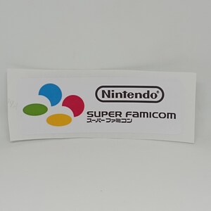 Puede incluir: Una pegatina blanca con el logotipo de Nintendo y el texto "SUPER FAMICOM" en inglés y japonés. El logotipo de Nintendo está en un óvalo negro, y el texto Super Famicom está debajo.