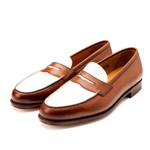 May include: A pair of brown leather loafers with white leather accents. The loafers have a penny strap detail and a rounded toe.