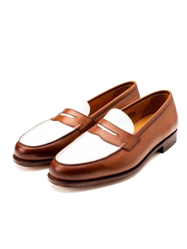 May include: A pair of brown leather loafers with white leather accents. The loafers have a penny strap detail and a rounded toe.