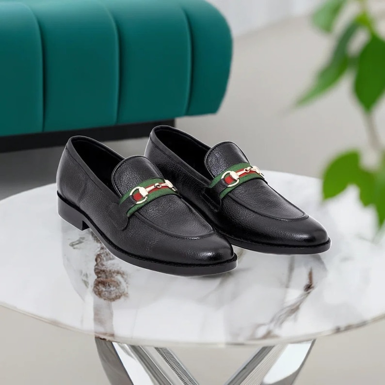 K&ouml;nnte beinhalten: Ein Paar schwarze Leder-Slipper mit einem gr&uuml;n-rot-wei&szlig; gemusterten Riemen &uuml;ber dem Oberteil. Die Slipper haben ein goldenes Schnallen-Detail am Riemen.