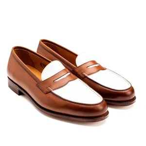 May include: A pair of brown and white leather loafers. The loafers have a white leather panel on the toe and a brown leather panel on the heel. The loafers have a penny strap detail.