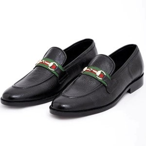 K&ouml;nnte beinhalten: Schwarze Leder-Slipper mit einem gr&uuml;n-rot-wei&szlig; gestreiften Band und goldenen Schnallen.