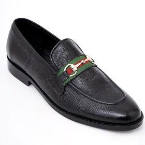 K&ouml;nnte beinhalten: Schwarze Leder-Slipper mit einem gr&uuml;n-rot-wei&szlig; gestreiften Band und einer goldenen Schnalle.