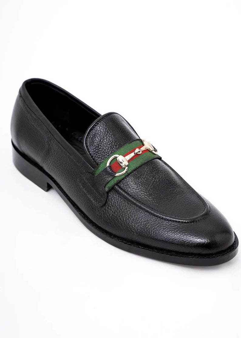 K&ouml;nnte beinhalten: Schwarze Leder-Slipper mit einem gr&uuml;n-rot-wei&szlig; gestreiften Band und einer goldenen Schnalle.