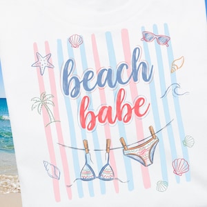 Op de afbeelding: Wit T-shirt met een strandthema. Het shirt heeft de woorden "beach babe" in blauw en rood script, samen met illustraties van schelpen, zonnebrillen en een bikini die aan een waslijn hangt. De achtergrond heeft blauwe en roze strepen.