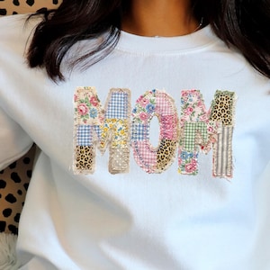 Peut inclure: Sweat-shirt blanc avec le mot "MOM" appliqué sur le devant. Les lettres sont faites de tissu patchwork à motifs variés, notamment floral, vichy et léopard. Un haut décontracté et confortable.