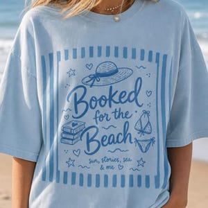 May include: Light blue t-shirt with a beach-themed graphic. The design includes a hat, bikini, books, and the words "Booked for the Beach" with "sun, stories, sea & me" below. The shirt has short sleeves and a relaxed fit.