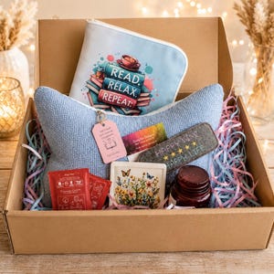 Könnte beinhalten: Eine Geschenkbox aus Karton mit einem hellblauen, strukturierten Kissen, einer Reißverschlusstasche mit dem Text READ. RELAX. REPEAT., einem Filz-Lesezeichen mit dem Text HAPPY MIND HAPPY LIFE, einem floralen Untersetzer, zwei Teepackungen und einem Glas.