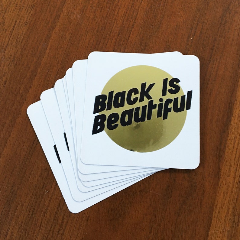 May include: A stack of white cards with a gold circle and the text "Black is Beautiful" in black letters.