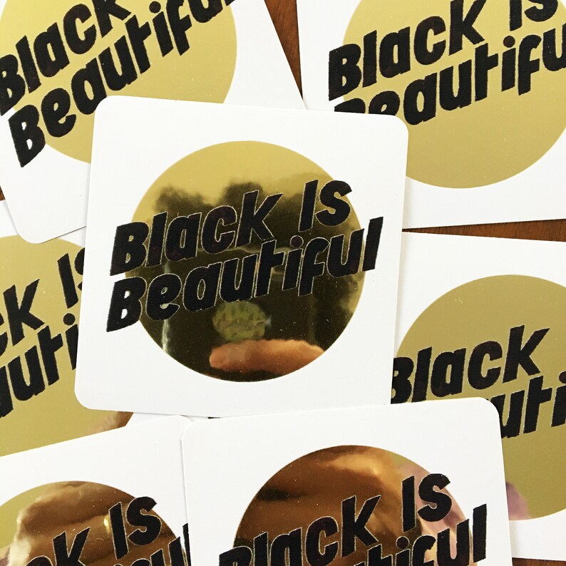 May include: Close-up of five gold stickers with the text "Black is Beautiful" printed in black. The stickers are on a wooden surface.