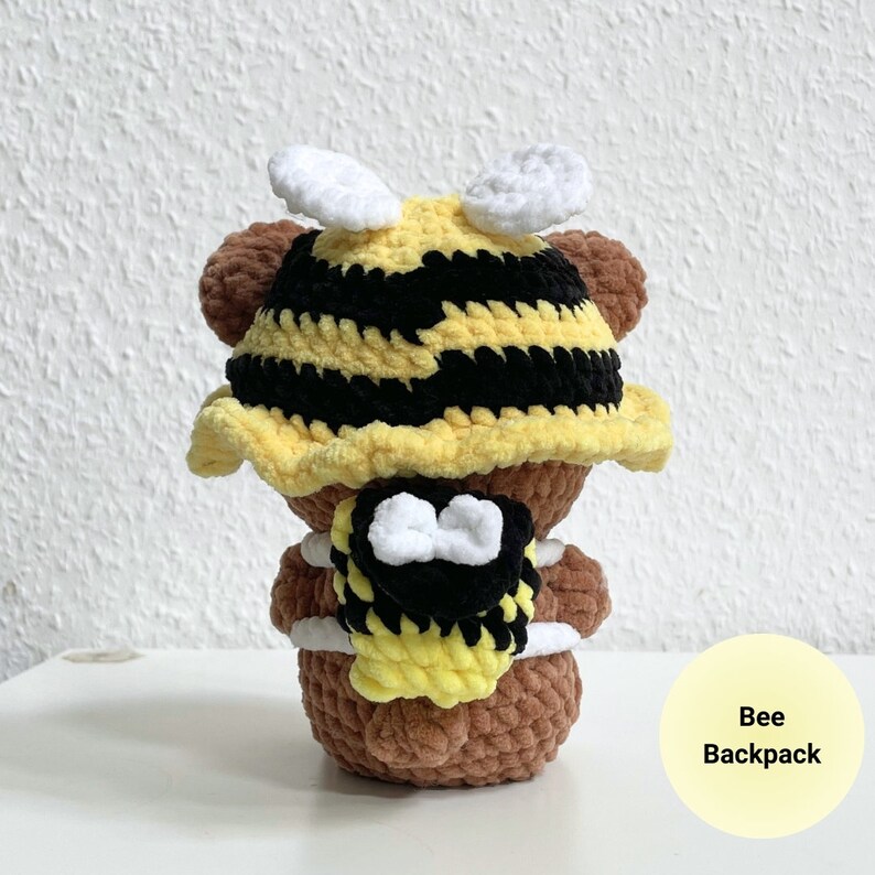 May include: A brown crocheted bear wearing a yellow and black bee-themed hat and backpack. The hat has white wings and the backpack has white accents.