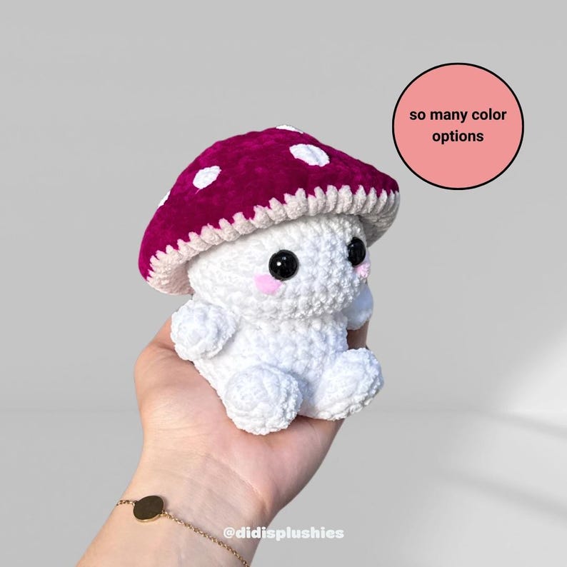 Op de afbeelding: Een handgemaakte gehaakte paddenstoel knuffel met een bordeauxrode kap en witte stippen. De paddenstoel heeft een wit lichaam, zwarte ogen en roze wangen. De tekst "so many color options" staat in een roze cirkel.