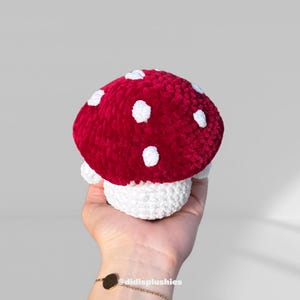 Op de afbeelding: Een handgemaakte pluche paddenstoel. De kap is rood met witte stippen en de steel is wit. Het speelgoed wordt in een hand gehouden. Een zacht, decoratief item.