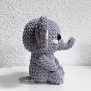 May include: A gray crocheted elephant plush toy with pink ears and black eyes. The elephant is sitting with its trunk raised.