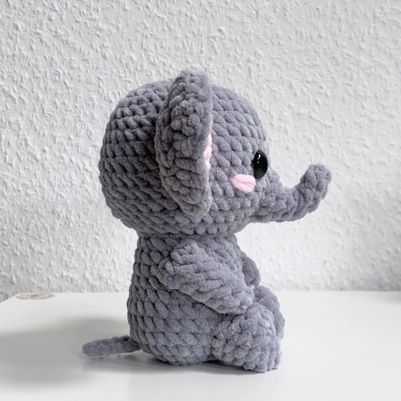 May include: A gray crocheted elephant plush toy with pink ears and black eyes. The elephant is sitting with its trunk raised.