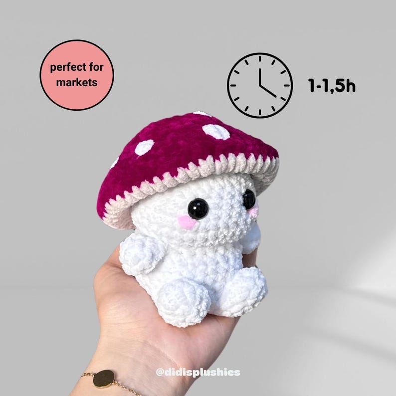 Op de afbeelding: Handgemaakte witte gehaakte paddenstoel knuffel met een bordeauxrode kap en witte stippen. De paddenstoel heeft zwarte ogen en roze wangen. De knuffel wordt in een hand gehouden. De tekst "@didisplushies" is zichtbaar.
