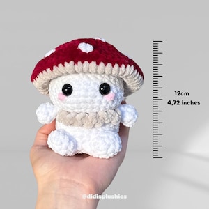 Op de afbeelding: Een handgemaakte gehaakte paddenstoel knuffel met een rode kap en witte stippen. De paddenstoel heeft een witte body, zwarte ogen en roze wangen. Hij is 12 cm hoog. De knuffel heeft een beige kraag.
