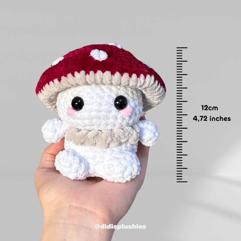 Op de afbeelding: Een handgemaakte gehaakte paddenstoel knuffel met een rode kap en witte stippen. De paddenstoel heeft een witte body, zwarte ogen en roze wangen. Hij is 12 cm hoog. De knuffel heeft een beige kraag.