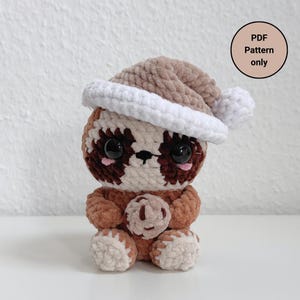 Puede incluir: Un perezoso de crochet con un gorro de Papá Noel marrón y blanco. El perezoso está sosteniendo una galleta de crochet marrón. La imagen incluye el texto "PDF Pattern only".