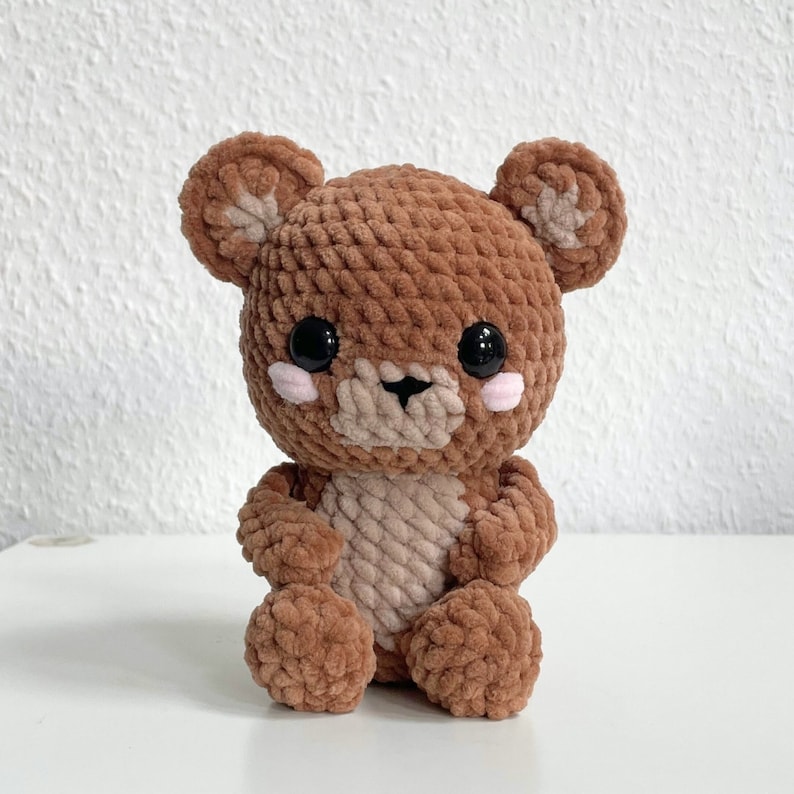 May include: A brown crocheted teddy bear with black eyes and pink cheeks. The bear has a soft, plush texture.