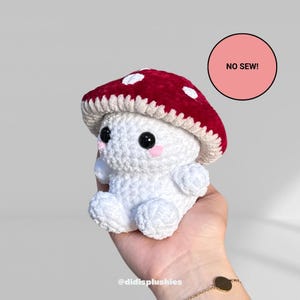 Op de afbeelding: Een gehaakte paddenstoel knuffel met een rode hoed met witte stippen. De paddenstoel heeft een witte body, zwarte ogen en roze wangen. De tekst "NO SEW!" staat in een roze cirkel. De knuffel wordt in een hand gehouden.