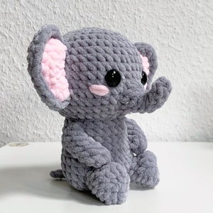 May include: A gray crocheted elephant plush toy with pink ears and black eyes. The elephant is sitting with its legs crossed.