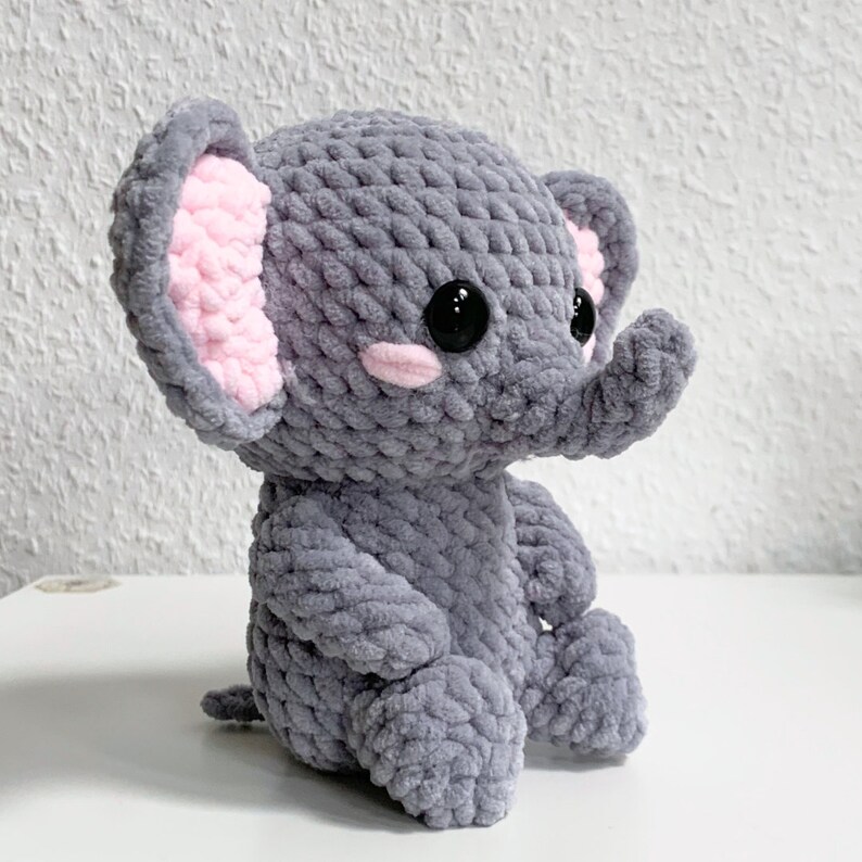 May include: A gray crocheted elephant plush toy with pink ears and black eyes. The elephant is sitting with its legs crossed.