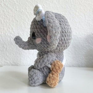 May include: A gray crocheted elephant plush toy with a white bow and a brown crocheted tail. The elephant is sitting on a white surface.