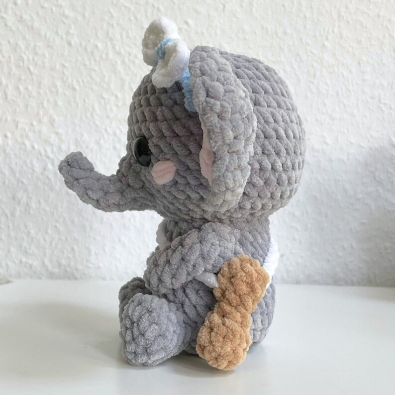 May include: A gray crocheted elephant plush toy with a white bow and a brown crocheted tail. The elephant is sitting on a white surface.